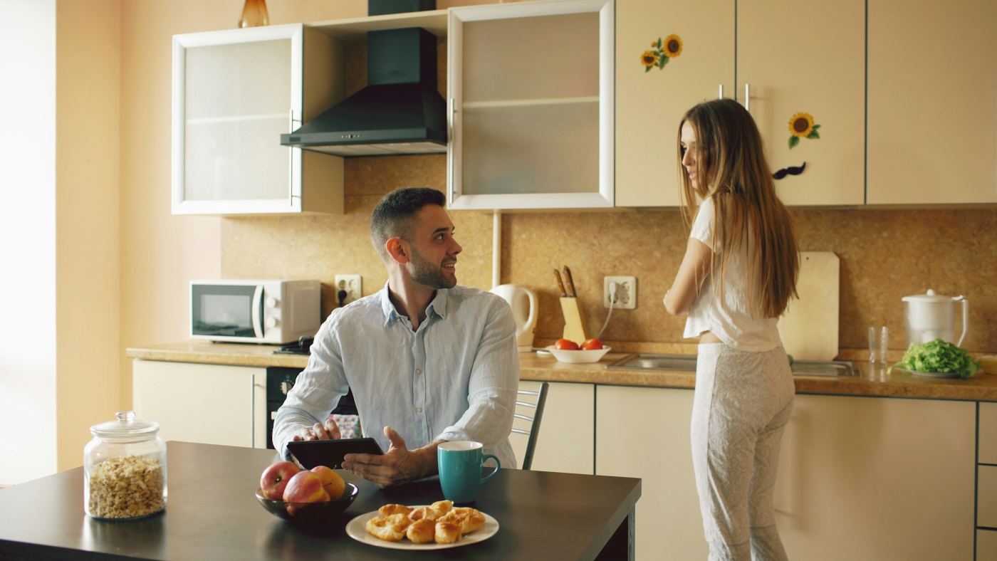 Een man met een tablet aan de keukentafel, in gesprek met zijn partner die aan het koken is.